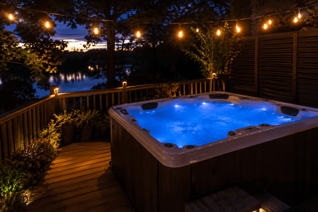 a well-lit hot tub on a wooden deck at dusk with string lights overhead and blue underwater lighting